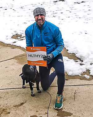 Justin posing with his dog while wearing running gear. Snow on grass, but not sidewalk.